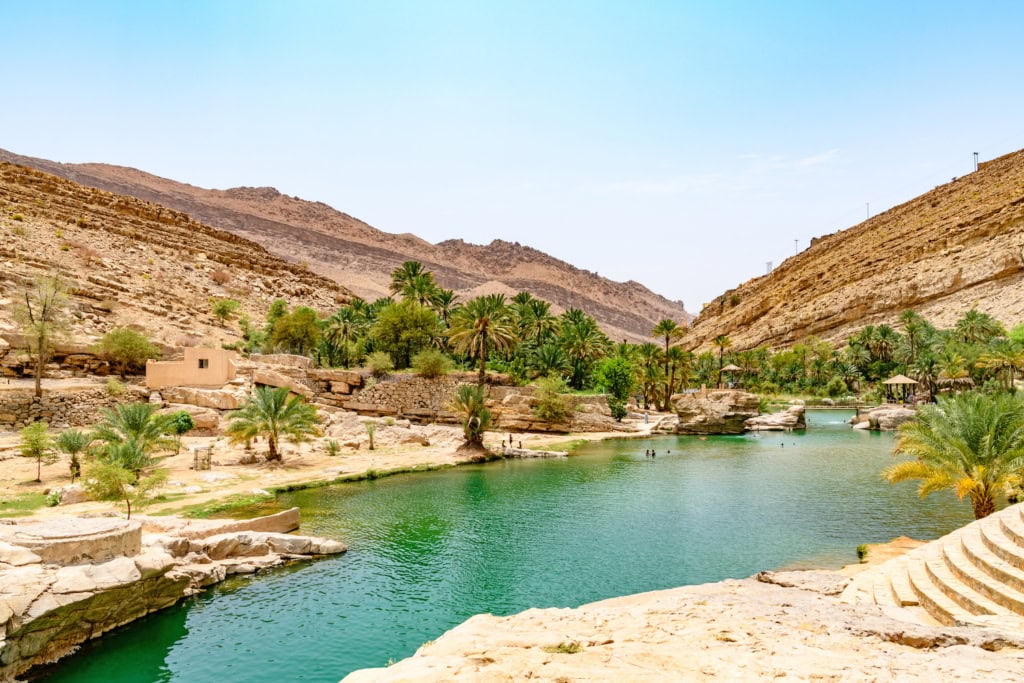 Oase im Wadi Bani Kalid im Oman: Türkisfarbenes Wasser, gesäumt von Palmen in brauner Wüstenlandschaft mit Bergen im HIntergrund.