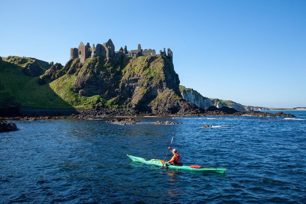 Ein Mann paddelt in einem Kayak an einer grünen Küste. Oben auf den Klippen thront die Ruine einer mächtigen Burg.