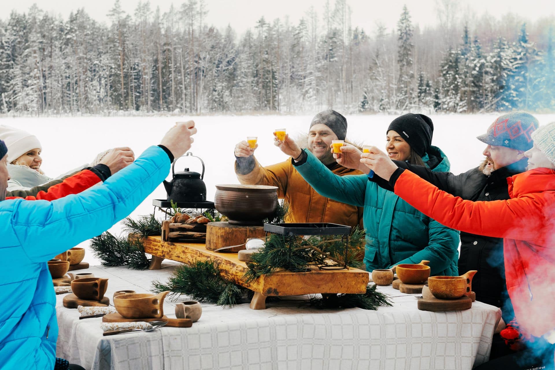 Gruppe von Menschen prosten sich in verschneiter Landschaft an einem Tisch sitzend zu. Auf dem Tisch stehen dampfende Schüsseln mit Essen.