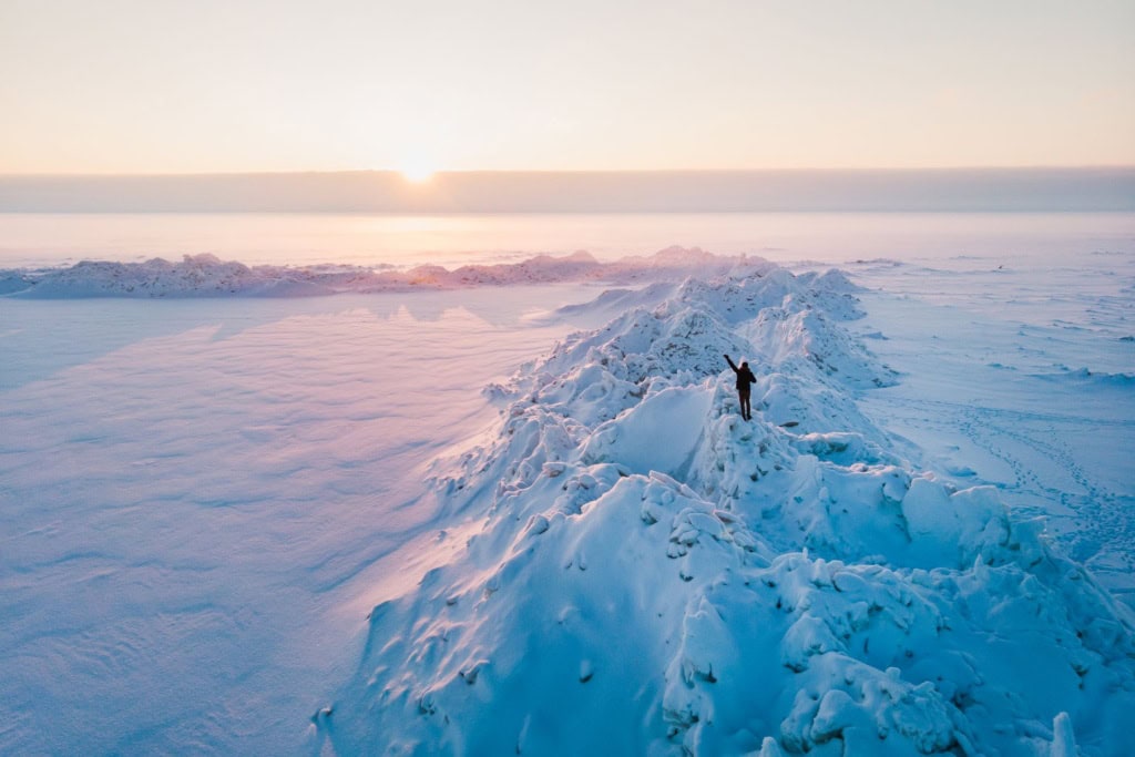 Verschneite Landschaft im Sonnenuntergang an einem See in Estland. Auf einem verschneiten großen Felsen steht ein Mann und streckt die Hände in den Himmel.