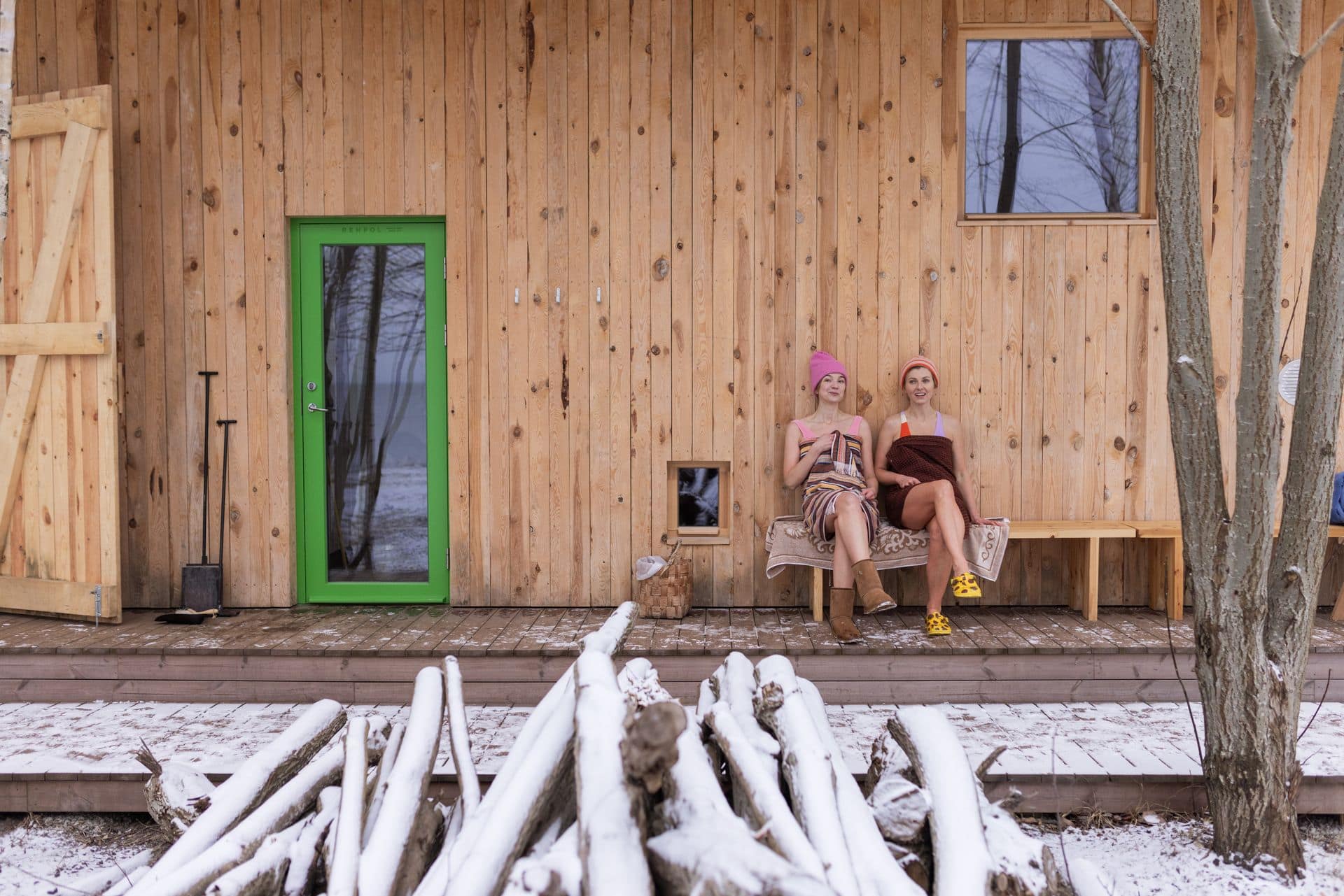 Zwei Frauen in Handtüchern sitzen vor einer Sauna in Estland , im Vordergrund Holz mit Schnee bedeckt.