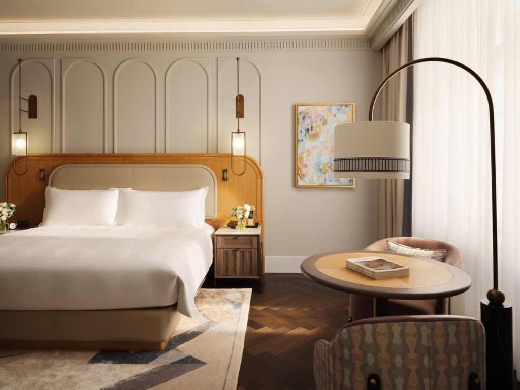Zimmer in einem Luxushotel; zur Linken ein breites Doppelbett mit breitem, hölzernen Kopfteil auf einem grauen Teppich. Der Boden ist mit dunklem Parkett verkleidet. Zur rechten vor einem Fenster zwei elegante Polstersessel an einem runden Beistelltisch, darüber eine geschwungene Stehlampe. Zur rechten kommt durch ein Fenster hinter einem weißen Vorhang Tageslicht herein.