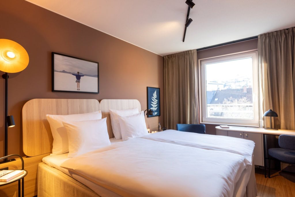 Doppelbett mit weißen Bezügen in einem Hotelzimmer; die Wand hinter dem Bett ist hellbraun gestrichen, zwei kleine Bilder hängen daran. Rechts vom Bett lässt ein großes Fenster Licht herein. Am Fenster steht eine schmaler Schreibtisch, links und rechts daneben zwei blaue Polsterstühle.