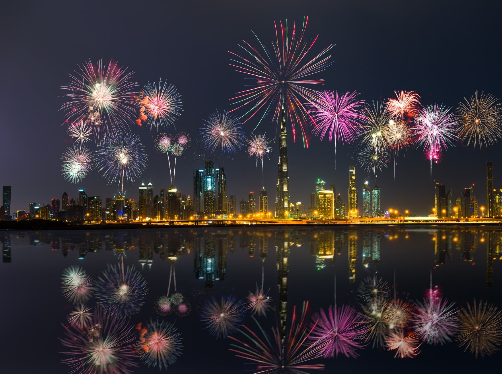 Die Skyline in Dubau mit mehreren Hochhäusern, darunter das Burj Khalifa; darüber steigt ein kunterbuntes Feuerwerk auf, das sich im Wasser im Vordergrund spiegelt.