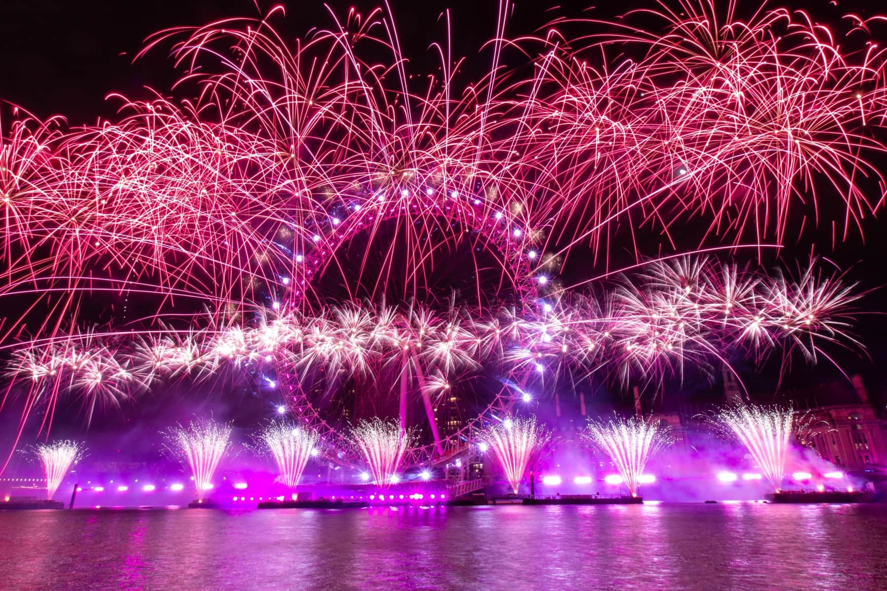 Vor dem Riesenrad "London Eye" steigt ein gigantisches Feuerwerk in den nächtlichen Himmel, das die Themse im Vordergrund in ein pink-violettes Licht taucht.