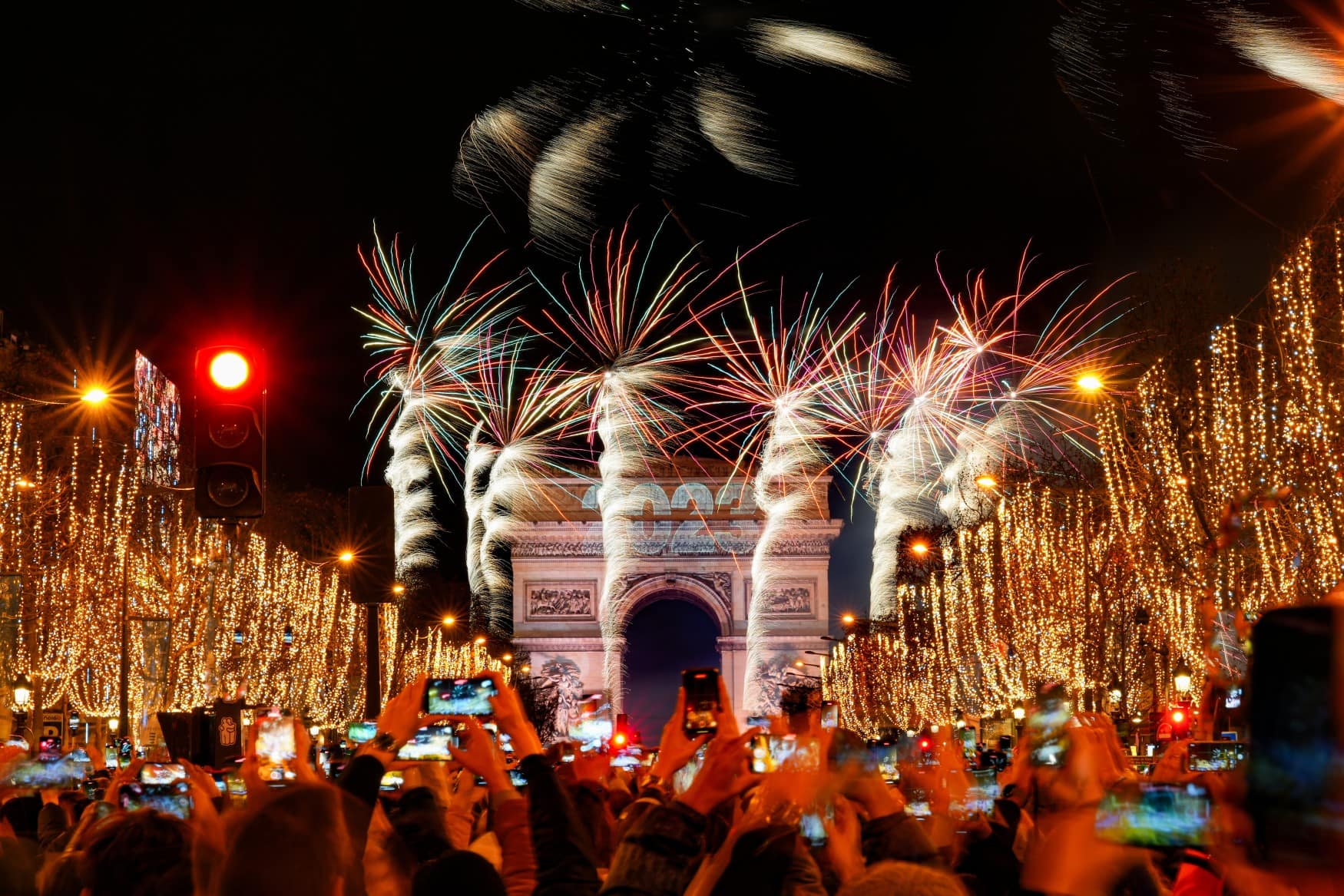 Eine große Menschenmenge blickt auf der gefüllten und festlich beleuchteten Champs Élysées in Paris auf dem Triumphbogen, über dem eine Feuerwerk in den Himmel steigt. Auf dem Bogen selbst wird die Jahreszahl "2023" projiziert.