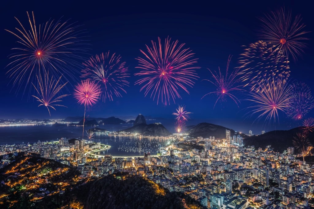 Luftaufnahme von Rio de Janeiro bei Nacht; über der hell erleuchteten Statt mit dem Zuckerhut im Hintergrund steigt ein riesiges Feuerwerk auf.