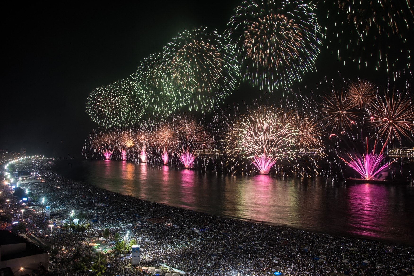 Luftaufnahme der Silvesterfeierlichkeiten an der Copacabana in Rio de Janeiro; der ganze Strand ist gefüllt mit Zuschauern, von mehreren Schiffen in der Bucht steigt ein choreographiertes Feuerwerk auf.
