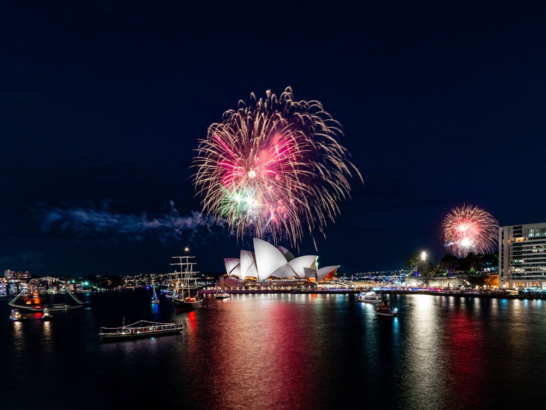 Über dem Opernhaus im Hafen von Sydney explodiert ein riesiges Feuerwerk. Im Vordergrund schwimmen einige Schiffe mit festlicher Beleuchtung.