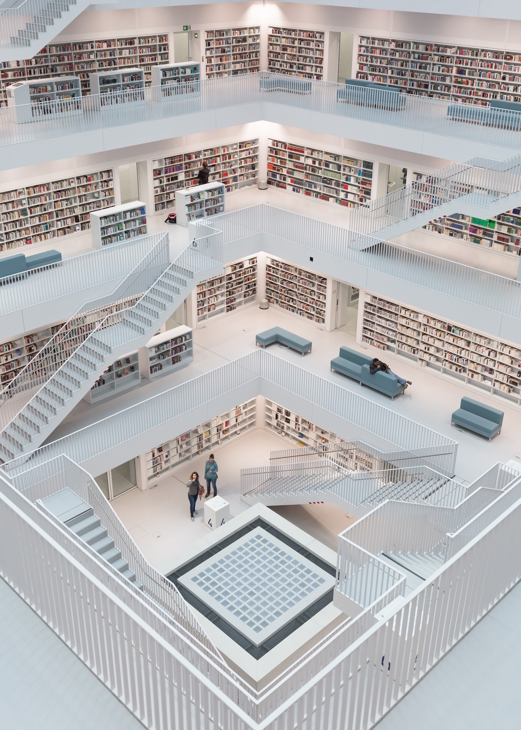 Blick aus einem der oberen Stockwerke ins Atrium der Stuttgarter Stadtbibliothek; die Ebenen rund ums quadratische Ebenen sind alle weiß, ebenso die Regale, die Böden und die Treppen. Die bunten Bücherrücken und die Menschen in der Bibliothek heben sich dadurch ab.