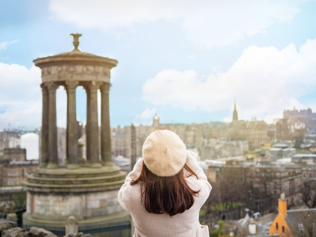 Frau von hinten mit dunklen Haaren, weißer Baskenmütze und weissem Mantel fotografiert in Edinburgh die Stadt von einem Hügel aus. Im Vordergrund das Dugald Stewart Monument auf dem Carlton Hill.