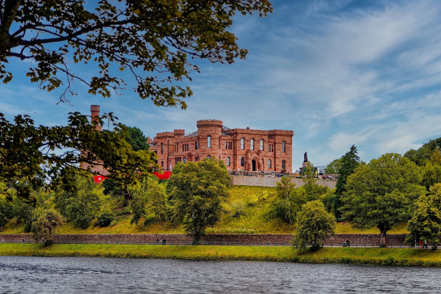 Blick auf das Inverness Castle im Sommer. Das Schloss liegt auf einer Anhöhe und ist eingerahmt von grünen Bäumen. Im Vordergrund ist ein Fluss.