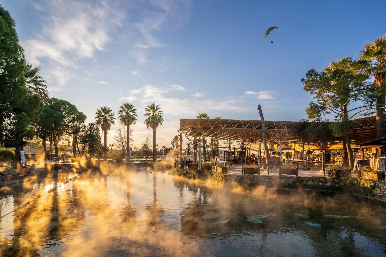 Thermalbad in Pamukkale in der Türkei im Abendlicht, ein großes, dampfendes Naturbecken, am Horizont Palmen und auf der rechten Seite befindet sich ein überdachter öffentlicher Bereich mit Tischen und Stühlen zum Ausruhen.