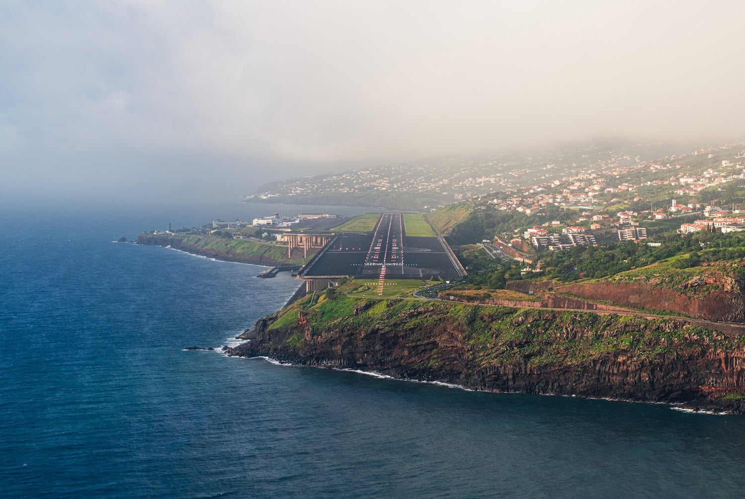 Madeira Landebahn direkt an der Küste der Insel, auf der linken Seite das Meer, auf der rechten das Land mit Häusern und grüner Landschaft.
