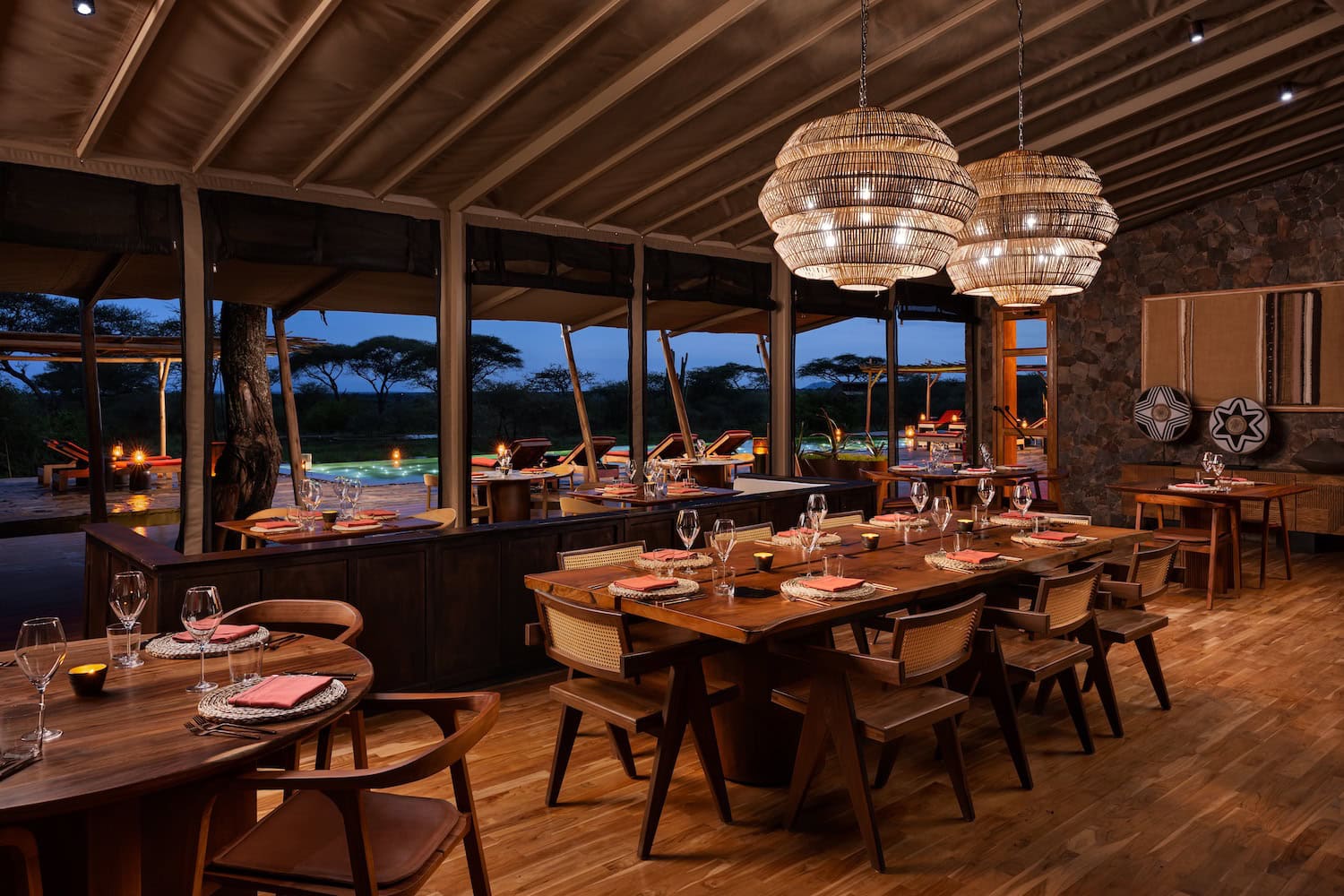 Restaurant mit offenen Bereich am Abend, dunkles Holz, dämmriges Licht, im Hintergrund Bäume der afrikanischen Serengeti und ein Pool.