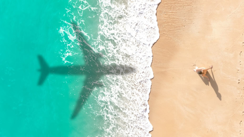 Schatten eines Flugzeugs über karibisch blauem Meer am Strand, Foto aus Vogelperspektive mit einer einzelnen Frau, die im Sand sitzt.