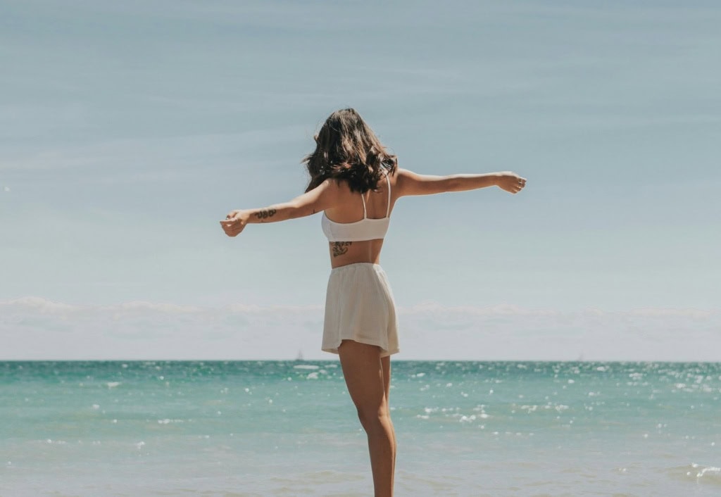 Eine junge Frau in Strandkleidung steht an einem Strand, den Rücken zum Betrachter und zum Meer hingewandt, und streckt beide Arme von sich.