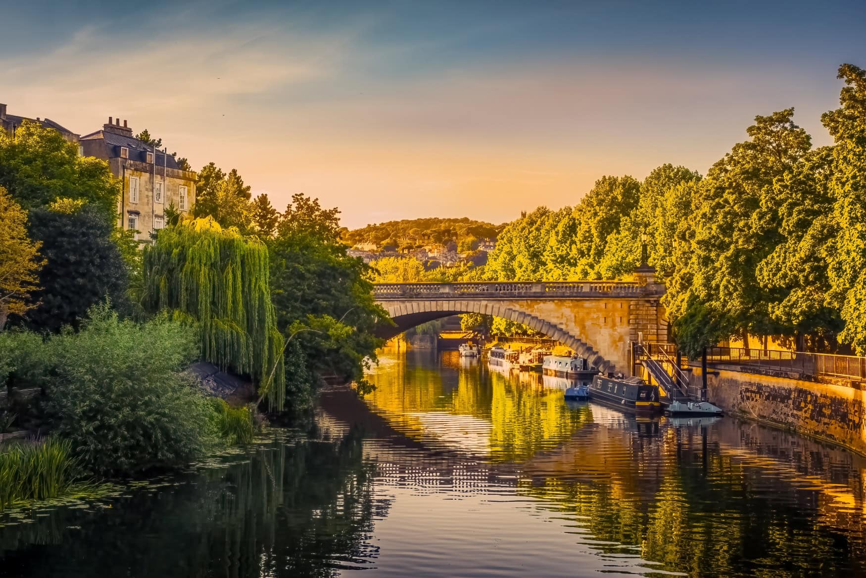 Über einen Fluss spannt eine Brücke aus fast weißem Stein. Das Flussufer ist zur Rechten befestigt, zur Linken von Bäumen begrenzt, zwischen denen ein zweigeschossiges, weißes Haus mit schwarzem Dach herausragt. Alles ist in das goldene Licht der untergehenden Sonne getaucht.
