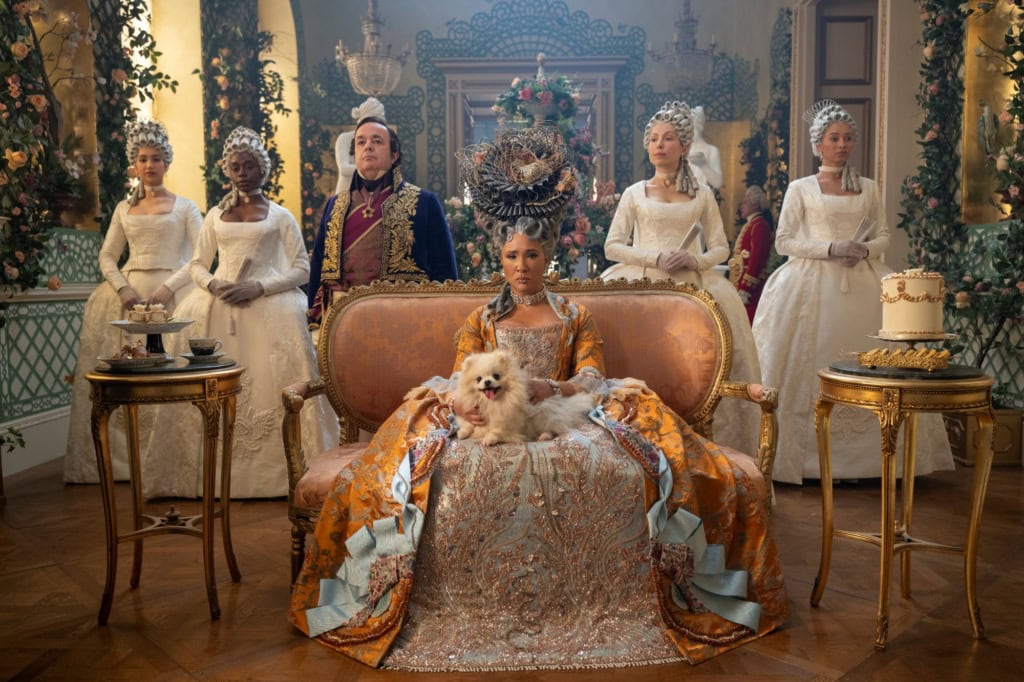 Still aus der Serie »Bridgerton«; Auf einem kleinen Sofa sitzt eine Frau in gold-silbernen Kleid mit einem weißen Hudn auf dem Schoß und blickt in die Kamera. Hinter ihr stehen aufgereiht vier Frauen in weißen Kleidern und ein Mann mit blau-goldenem Anzug.
