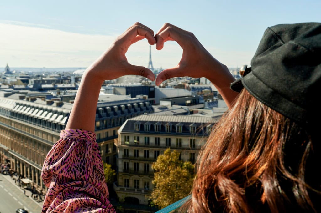 Eine Person hält ihre Hände zu einem Herzen zusammen, in dessen Mitte der Eiffelturm am Horizont über den Dächern von Paris sichtbar wird.