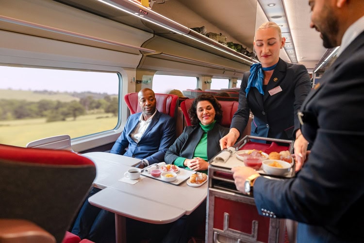 Zwei Menschen sitzen in einem Viererabteil mit Tisch des Eurostar. Zwei Mitarbeiter servieren ihnen Speisen von einem Servierwagen.