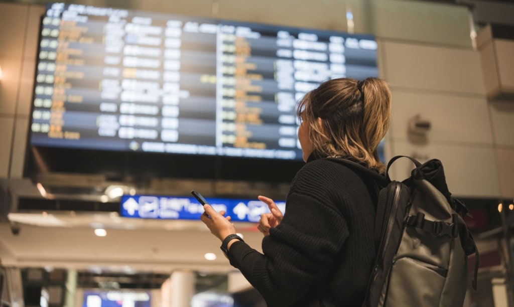 Eine Frau steht vor einem Abflugsterminal in einem Flughafen und gleicht die Daten mit ihrem Smartphone ab.