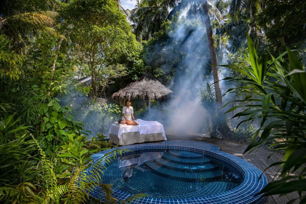 Inmitten einer Dschungellandschaft ist ein kleiner, runder Pool in eine Holzterrasse eingelassen. An deren Rand wird eine Person auf einem Tisch von einer anderen Person massiert.