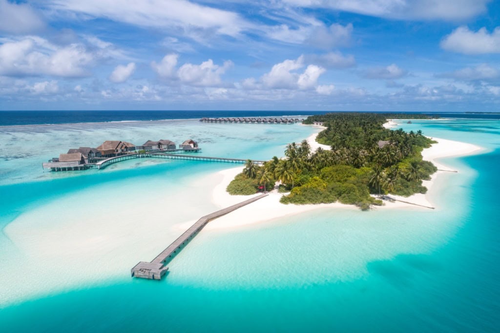 In mitten einer türkisblauen Lagune liegt eine langezogene, grüne Insel, von der links ein Steg abgeht, an dessen Ende mehrere Pavillons auf dem Wasser stehen.