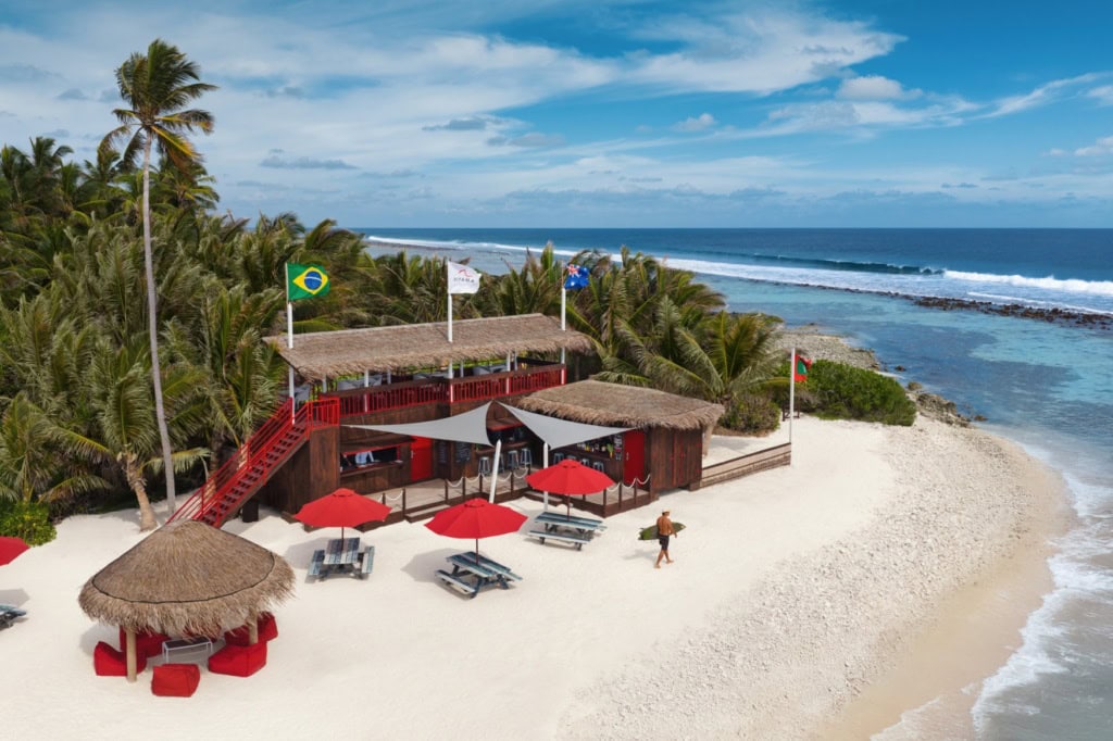 An einem tropischen Strandabschnitt steht eine zweigeschossiger Holzpavillon mit rotem Geländer am oberen Stockwerk. Dahinter ein kleiner Wald, davor einige Sitzgelegenheiten. Direkt am Strand geht ein Mann mit einem Surfbrett unterm Arm Richtung Wasser.