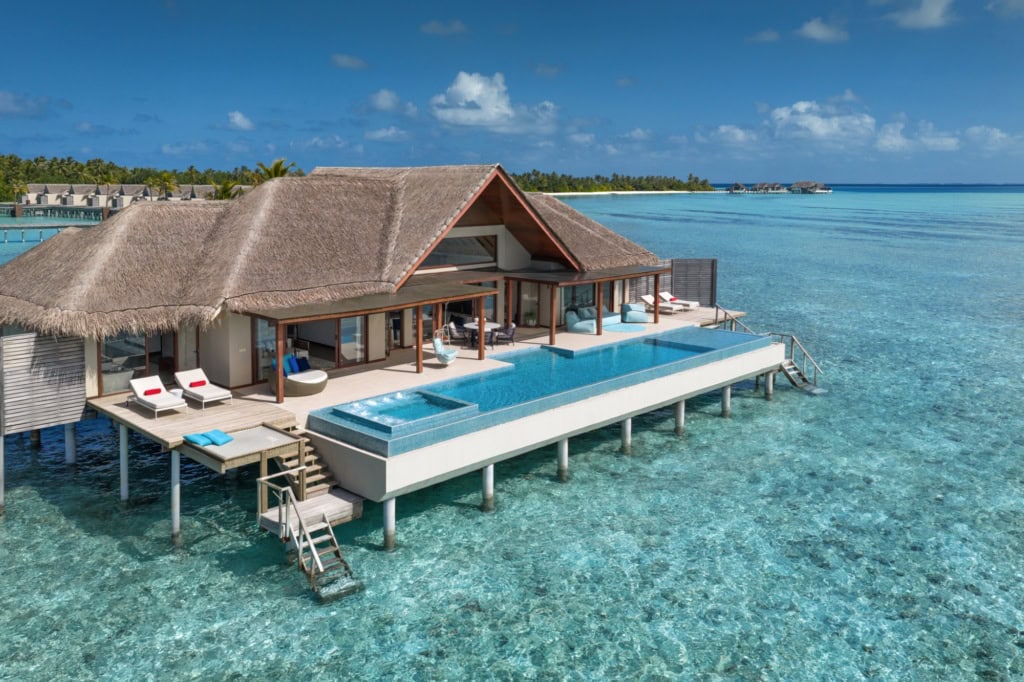 In einer türkisblauen Lagune steht ein eleganter Pavillon mit Strohdach und einem Pool auf Stelzen im Wasser.