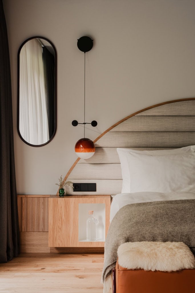 Blick auf das Bett in einem eleganten Hotelzimmer; rechts eine Bedhälfte, dahinter ein halbrundes, grau gepolstertes Kopfteil. Links ein hölzerner Nachttisch, darüber eine kugelrunde Lampe mit orangefarbenem Schirm, links dahinter ein ovaler, hochkanter Spiegel.