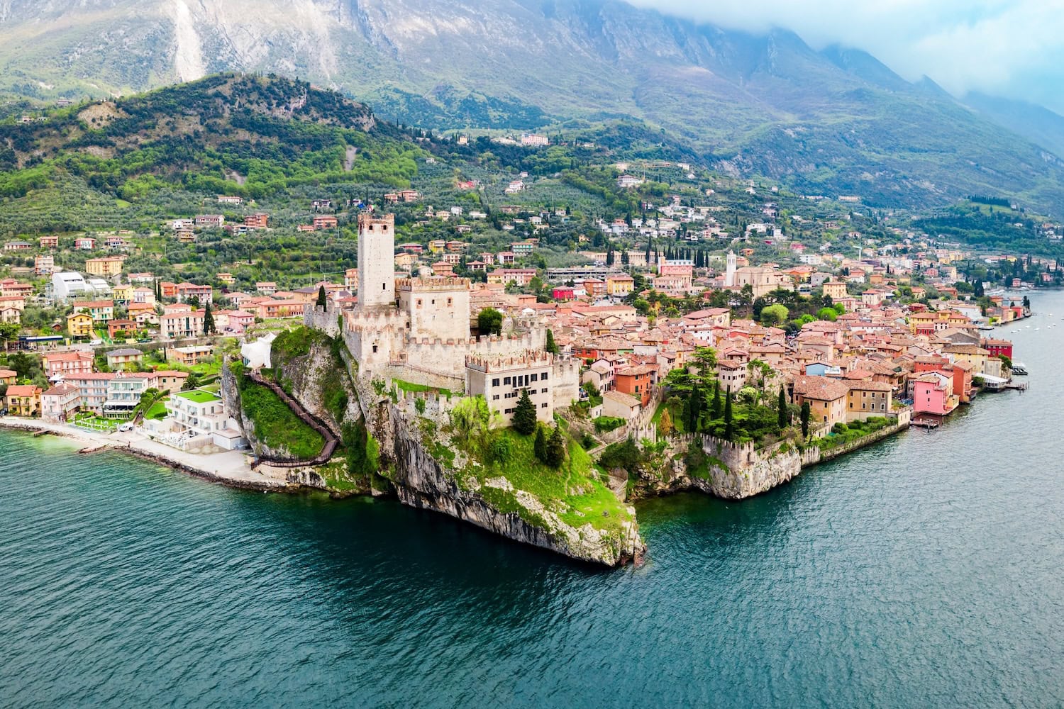 Luftaufnahme auf das Castello Scaligero am Gardasee, das auf einer kleinen Landzuge fast in den See hineinragt. Drumherum ist der kleine Ort, im Hintergrund hohe Berge.