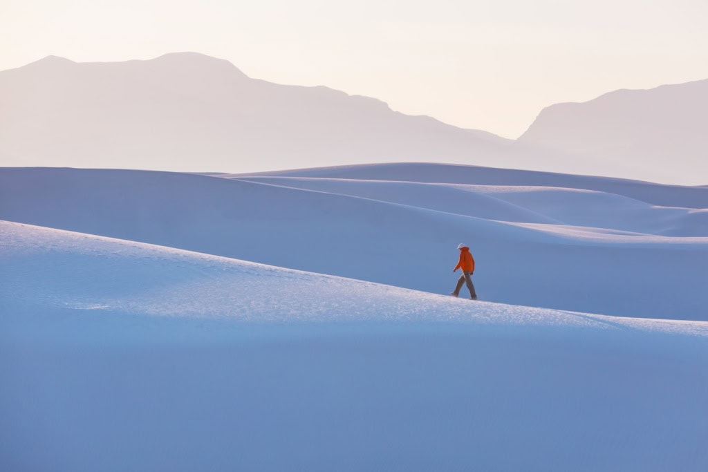 Ein einzelner Mann in roter Jacke, der inmitten der weiße Sanddünen vom Withe Sands Nationalpark spaziert, die im Licht fast blau wirken. Im Hintergrund sind die Umrisse von Felsen und Bergen zu erkennen, aber ganz blass und zart.