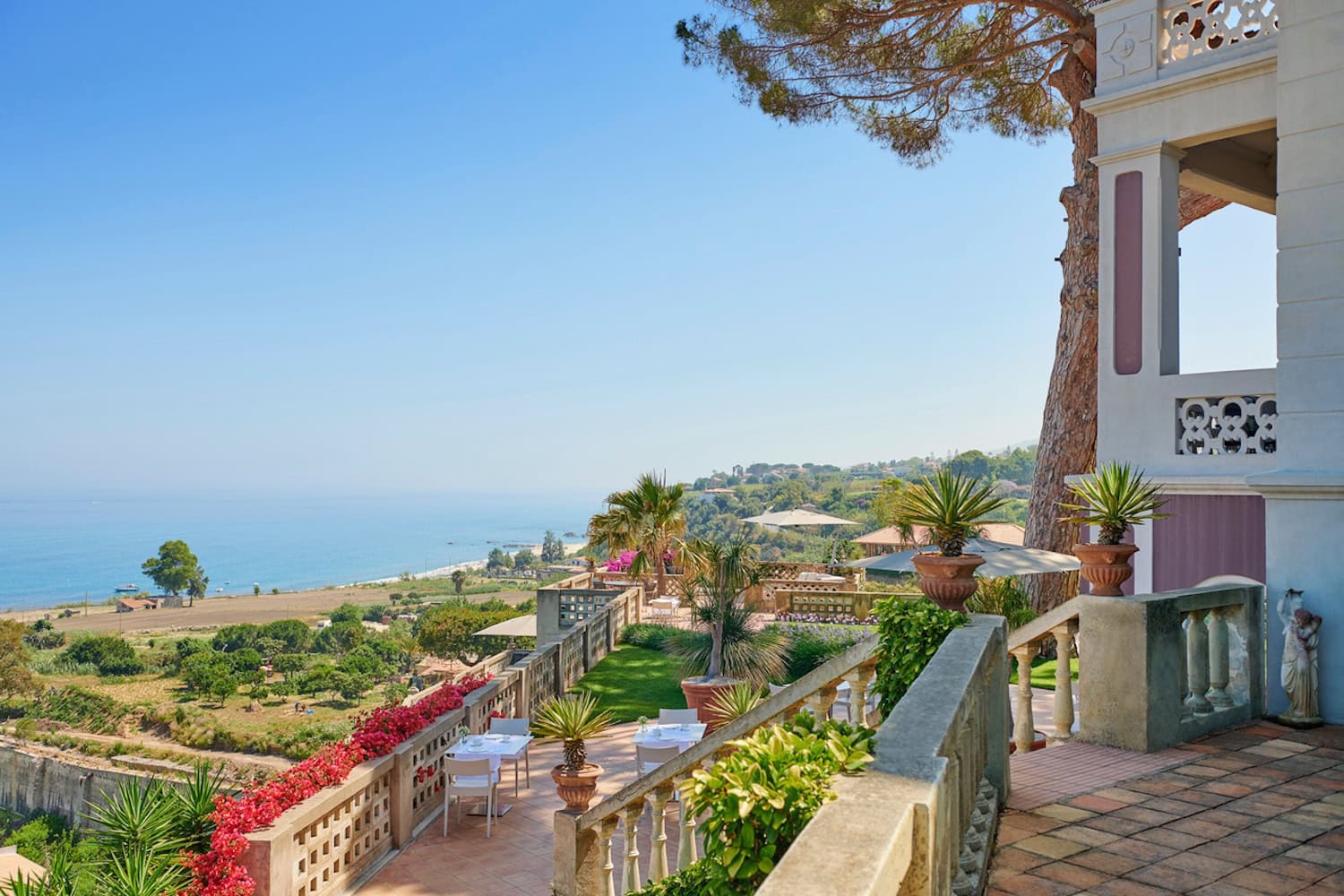 Blick von der Villa Paola auf die Küste und das Meer Kalabriens. Auf dem Bild ist die steinernde Terrasse der Villa zu sehen und ein Stück des Gartens mit grünem Rasen und Palmen. Auf der zweigeschossigen Terrasse stehen Tische und Stühle, darunter die Landschaft bis zum Meer mit Bäumen und Feldern.