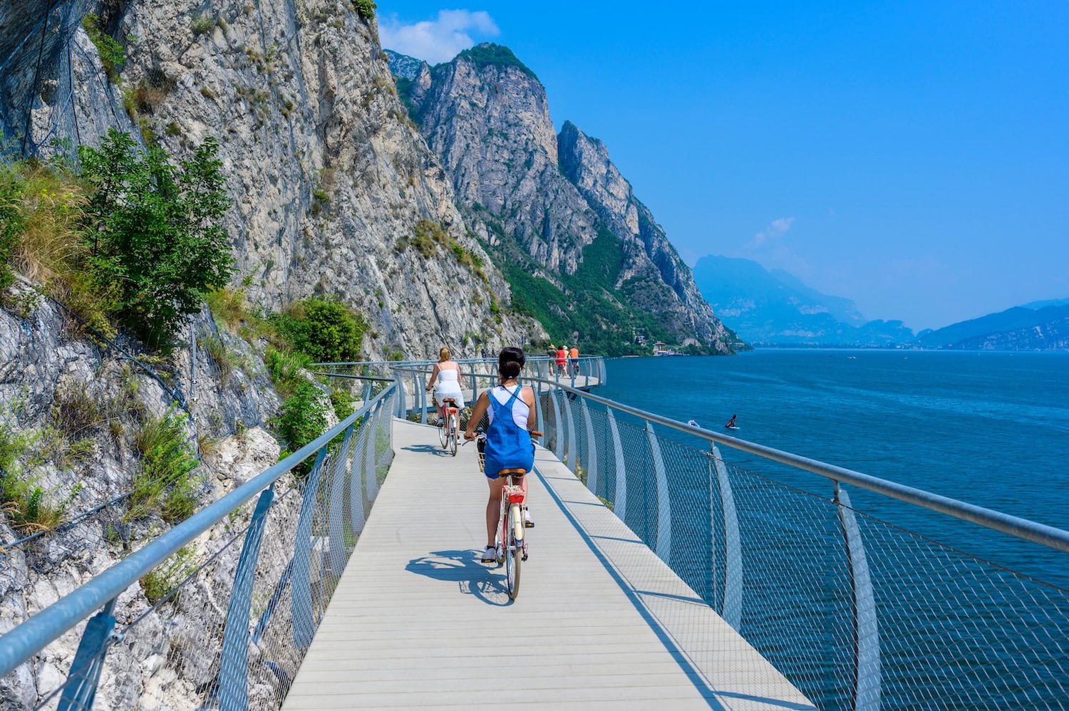 Zwei Radfahrerinnen im Sommer auf dem Limone del Garda, einem neuen Radweg mit Geländer entlang der steilen Berge des Gardasees. Links die Berge, rechts, der See, am Horizont weitere Berge.