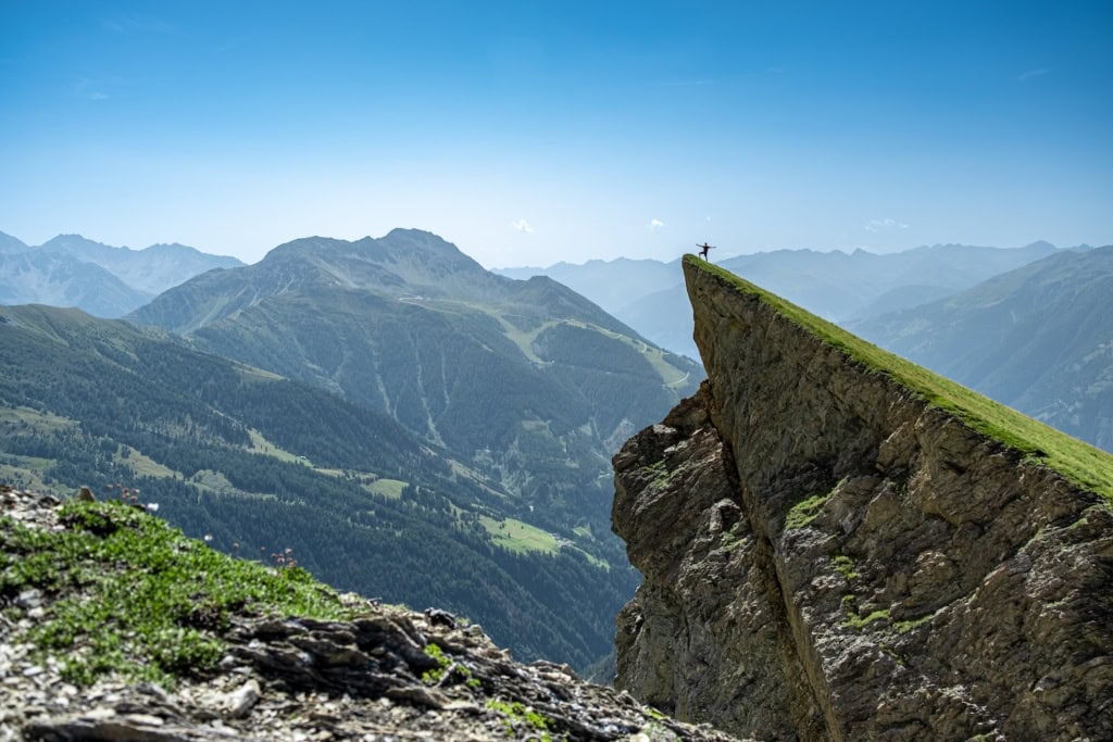 Panoramabild auf die Osttiroler Berge mit blauem Himmel: Auf einem markanten Berg, der spitz zur Seite ragt, steht auf dem Gipfel eine einzelne Person und reckt die Hände in den Himmel. Von der Fotoperspektive aus sieht sie sehr klein aus.