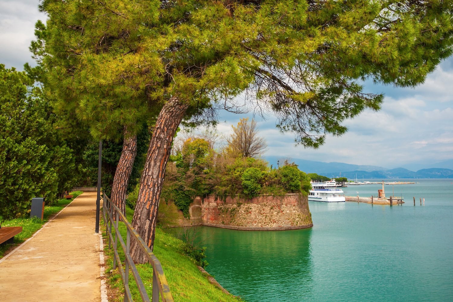 Peschiera del Garda, ein kleiner Ort am Gardasee, ein Weg mit grünen Nadelbäumen, rechts das türkisgrüne Wasser des Sees, auf dem in einiger Entfernung ein Ausflugsboot schwimmt.