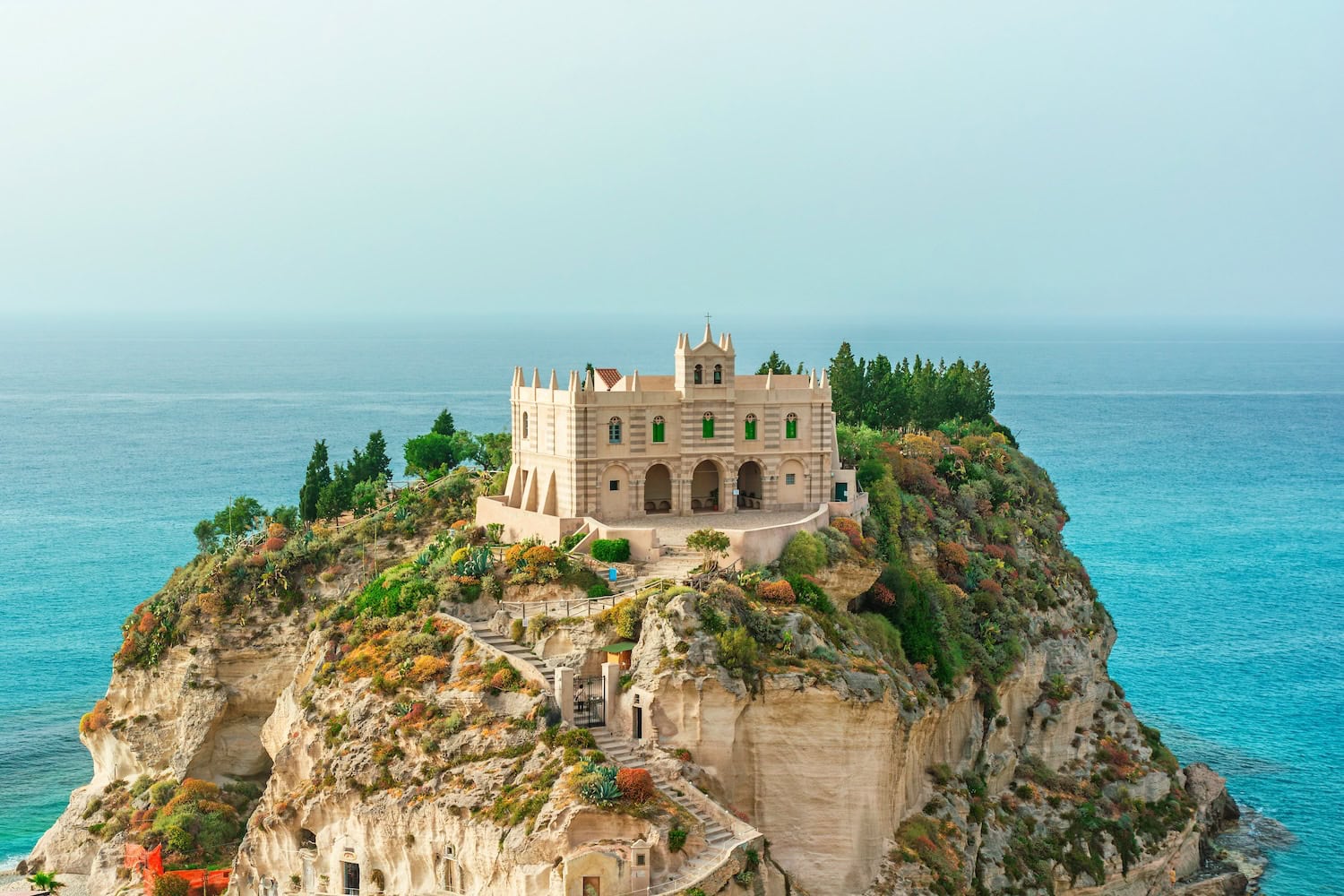 Blick auf die Kirche Maria dell'Isola auf einer hohen Klippe an der Küste Kalabriens im Ort Tropea. Die Kirche ist ganz oben, steile Treppen führen zu ihr hinauf. Im Hintergrund ist das türkisfarbene Meer zu sehen.