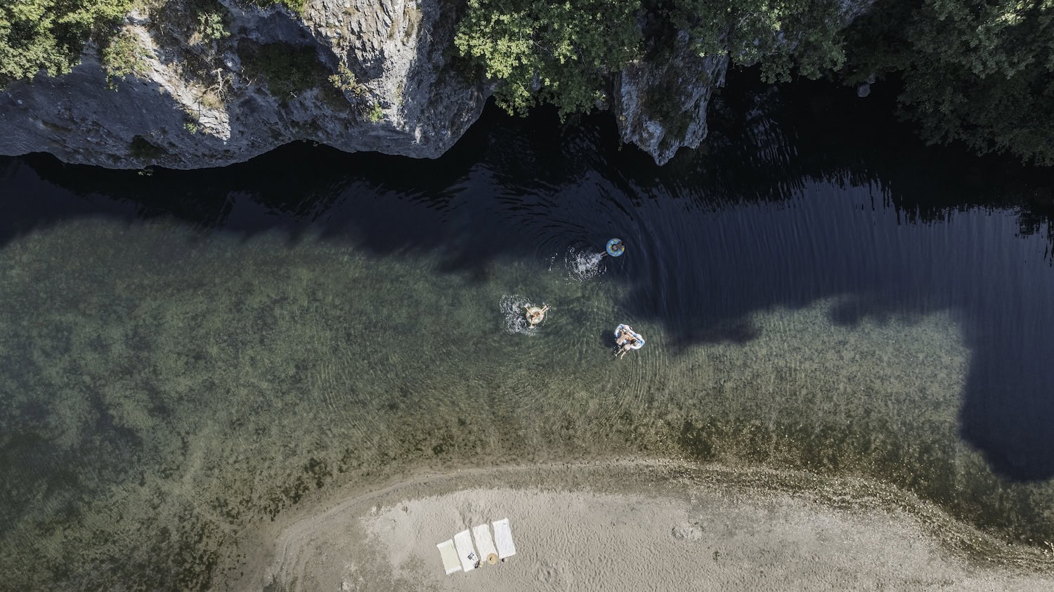 Luftaufnahme von Menschen, die in einem Fluss mit sehr klarem Wasser baden. Alle schwimmen auf bunten Schwimmreifen und sind nur ganz klein erkennbar. Am sandigen Flussufer liegen vier Handtücher ausgebreitet, am oberen Ende des Bildes sind hohe Felsen mit Sträuchern zu sehen.