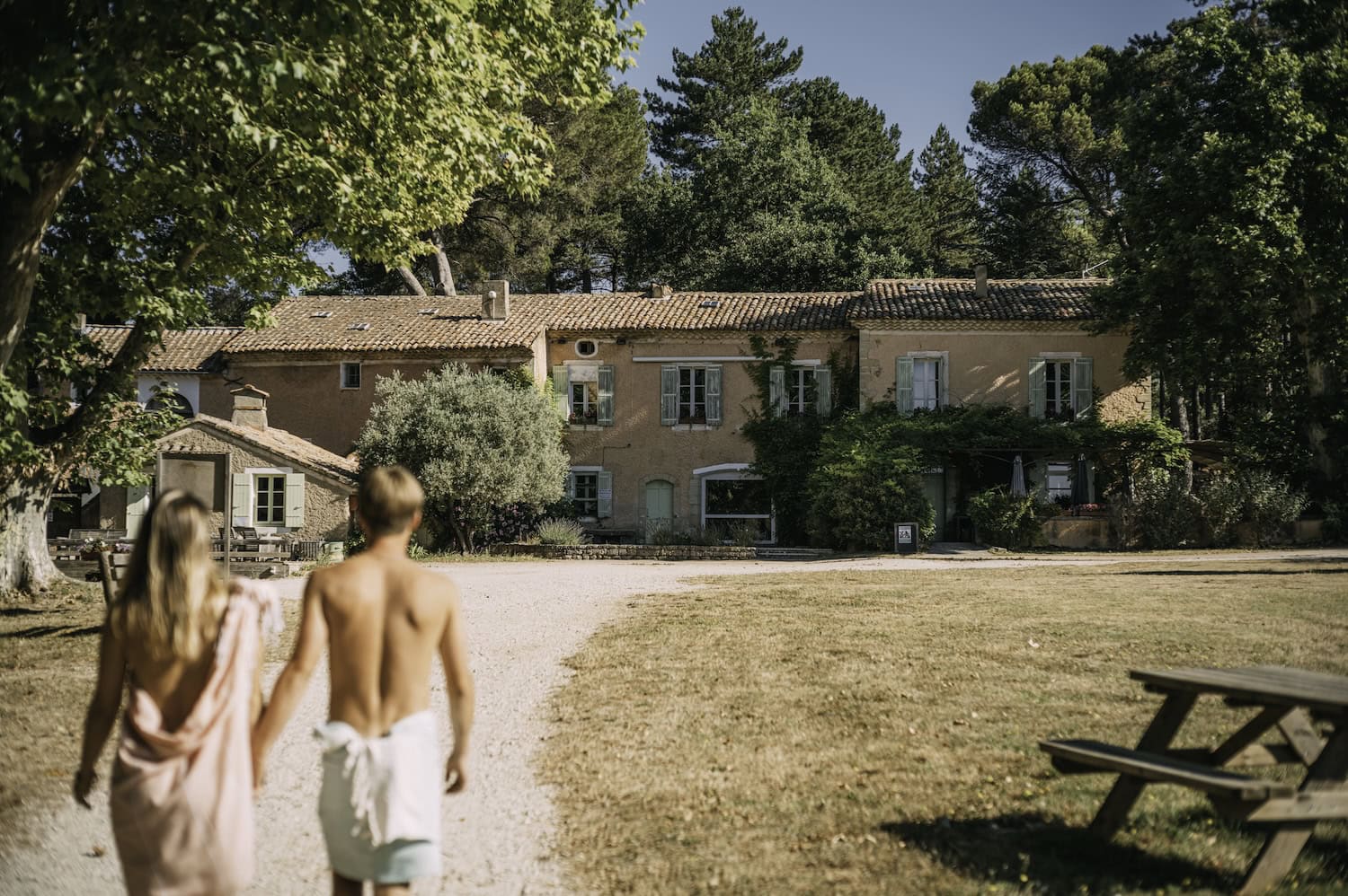 Ein französisches Landhaus aus braunen Stein mit türkisen Fensterläden inmitten von Bäumen auf einem FKK-Campingplatz im Sommer. Ein unscharf gestelltes Paar, in Tücher gehüllt, geht auf das Haus zu.