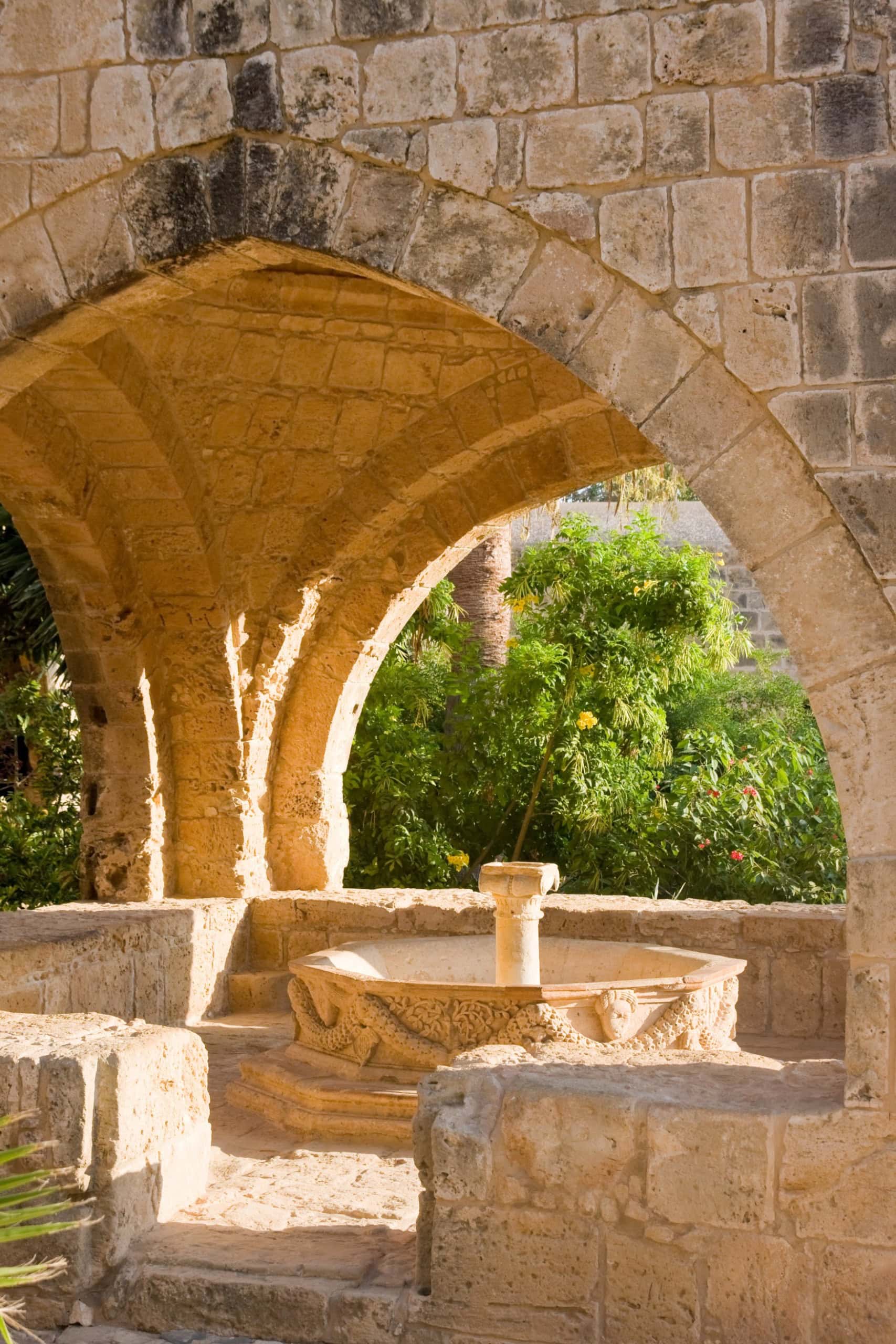 Ein gotischer Brunnen, überdacht von einem Gewölbe, mit offenen Wänden, durch die die Sonne hereinstrahlt.