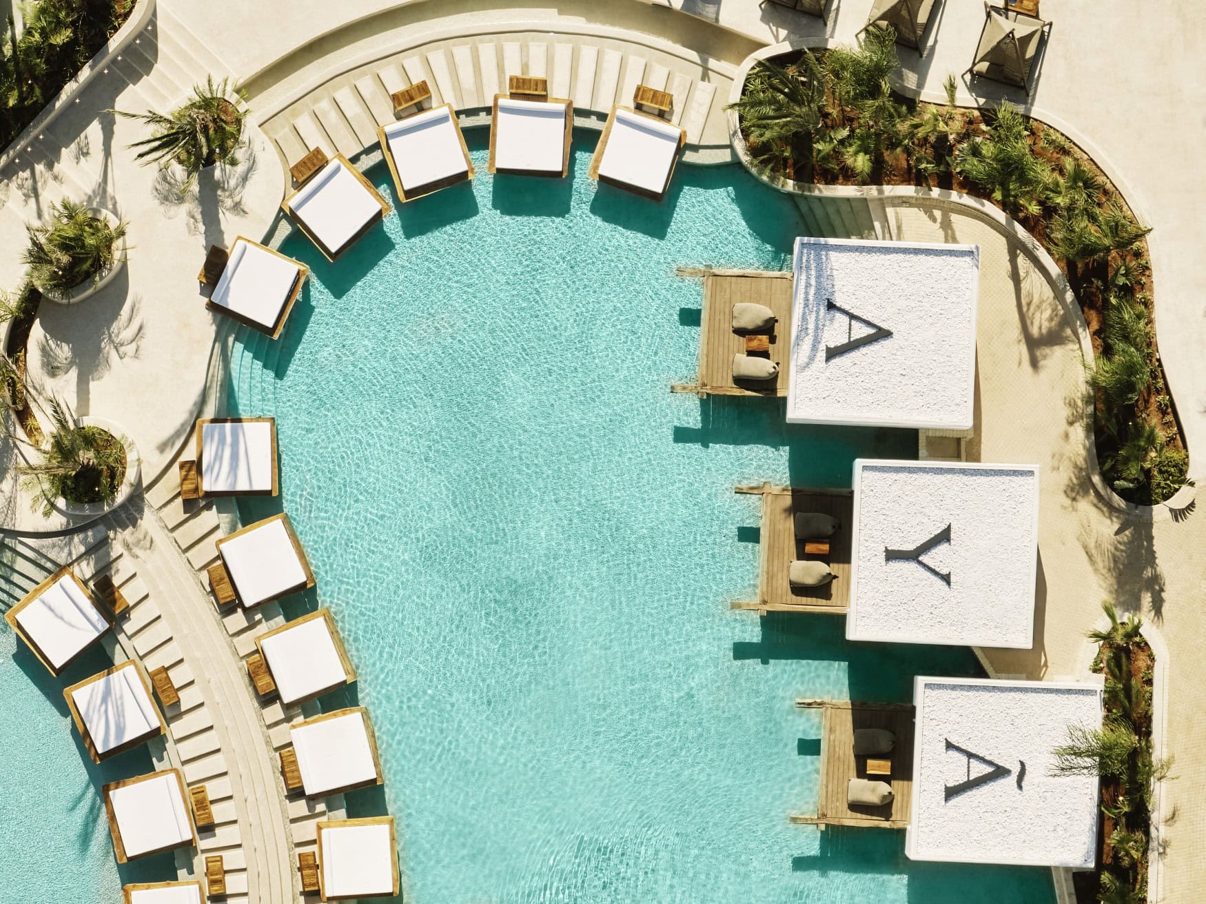 Luftaufnahme eines geschwungenen Pools in einem Resort auf Zypern. Am Rand ragen einige breite Liegen über die Wasseroberfläche. Auf der rechten Seite grenzen drei Bungalows an den Rand, auf deren Dächern die Buchstaben »A«, »Y« und »A« aufgebracht sind.