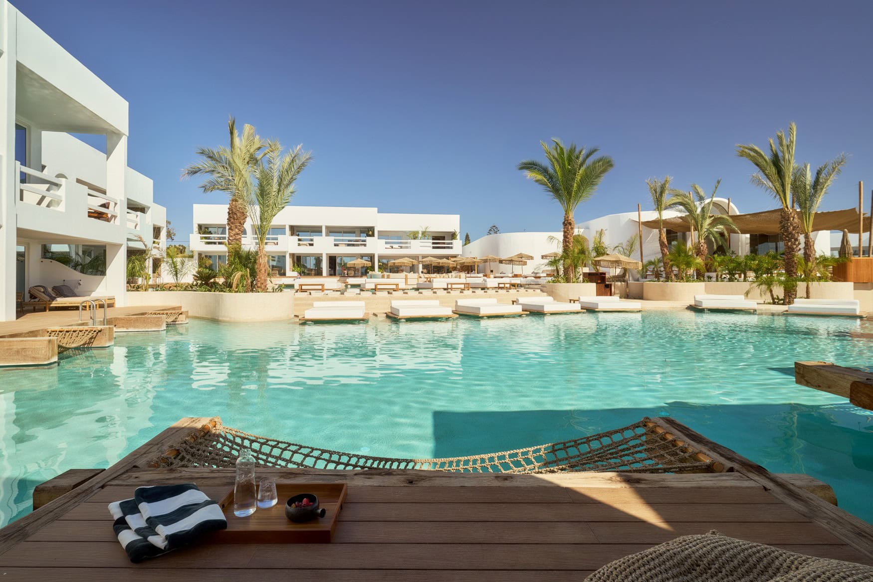 Blick von einem Liegedeck mit Hängematte auf einen breiten, türkis schimmernden Pool in einem Resort. Auf der anderen Seite des Pools ragen einige Polsterliegen über das Wasser.