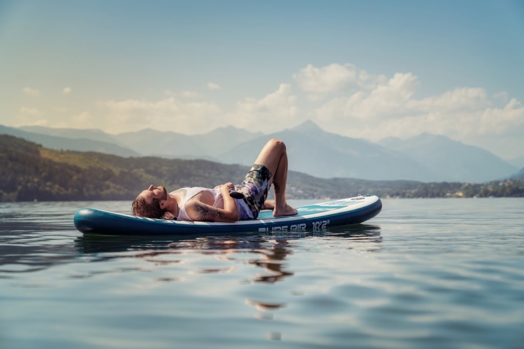 Mann liegt auf seinem Stand-Up-Paddle in der Sonne und entspannt auf dem Millstätter See im Sommer. Im Hintergrund Berge und weisse Wolken.