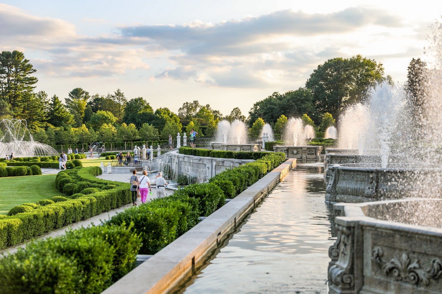 Wasserkanal und Springbrunnen in Longwood Gardens im warmen Abendlicht, Besucher spazieren entlang der Buchsbaumhecken
