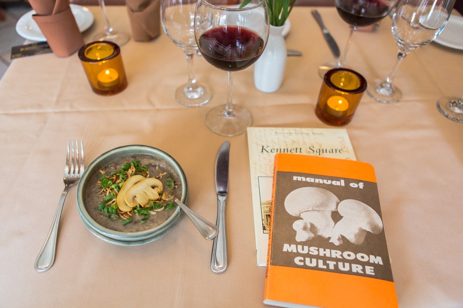 Gedeckter Restauranttisch in Kennett Square mit Champignon-Cremesuppe, Rotwein und dem Buch „Manual of Mushroom Culture"
