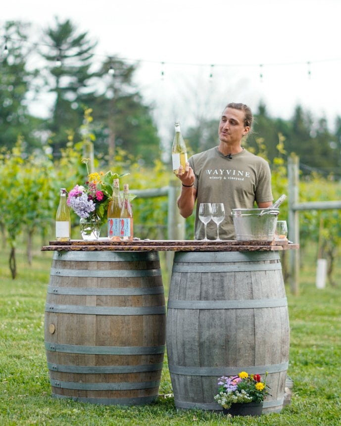 Winzer präsentiert eine Flasche Weißwein an Holzfässern im Außenbereich des Wayvine Winery & Vineyard