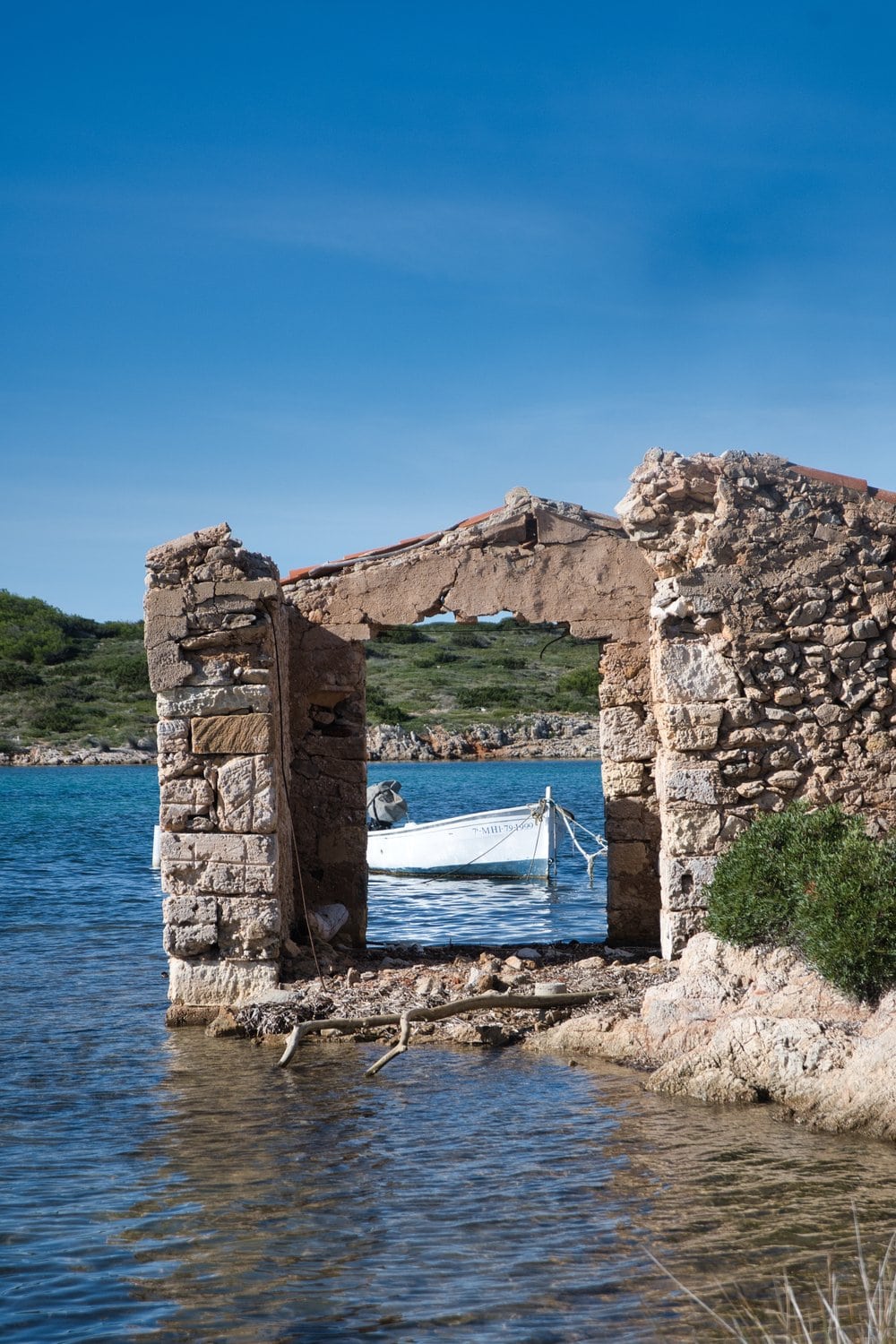 fischerboot-menorca.jpgVerfallene Steinmauern eines alten Bootshauses am Wasser auf Menorca, dahinter ein weiß-blaues Fischerboot vor strahlend blauem Himmel.