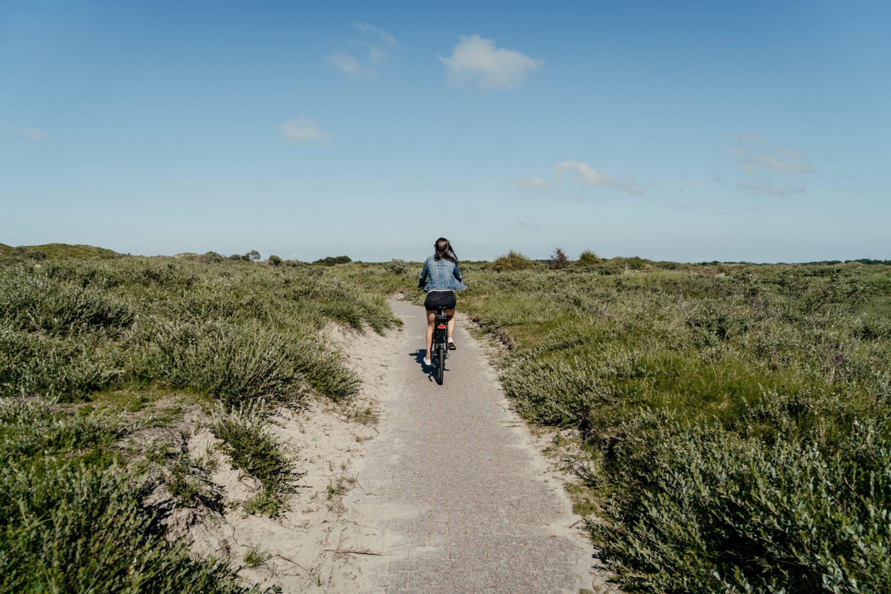 Eine Person fährt vom Betrachter weg auf einem Radweg, der durch grüne Dünen auf der Ostfriesischen Insel Borkum führt.