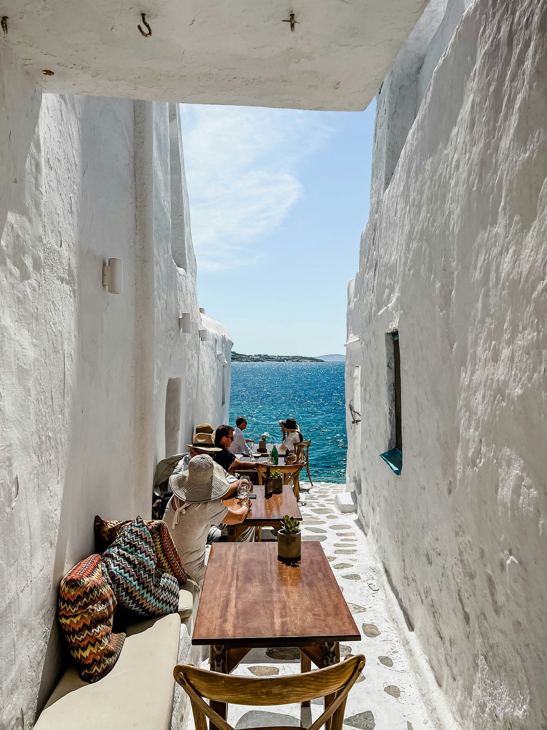 Cafégäste sitzen an einem Holztisch in einer schmalen weißen Gasse auf Mykonos mit direktem Blick auf das türkisblaue Meer